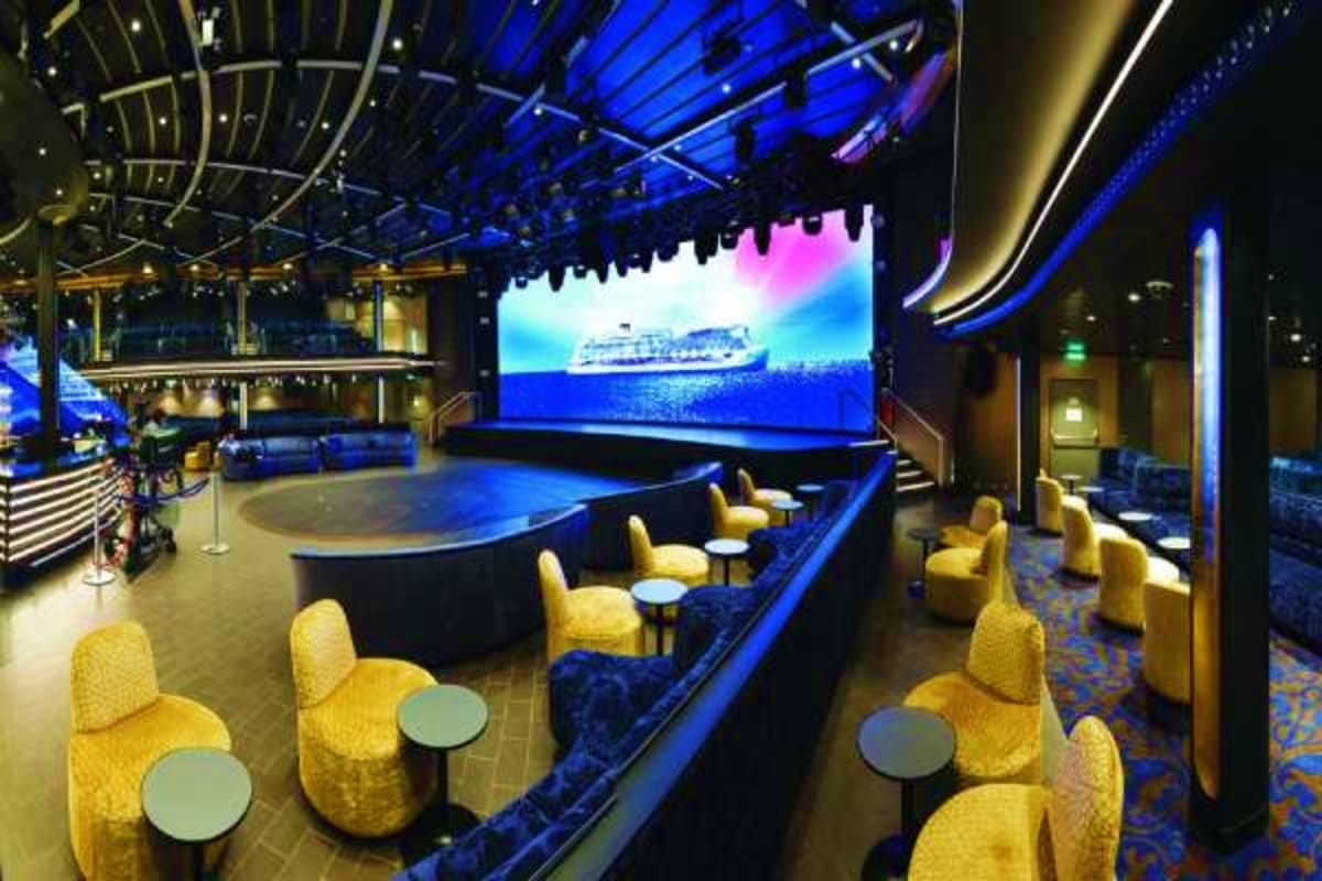 Modern cruise ship theater interior with yellow seats, blue ambient lighting, and large screen displaying a cruise ship at se